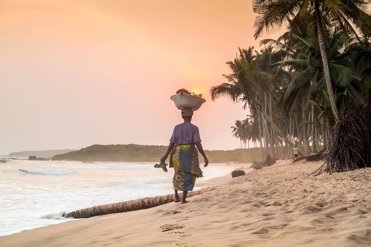 A serene, wide-angle shot of a woman seen from behind, walking along a sandy beach in Ghana at sunset. She is wearing a purple shirt and a colorful patterned wrap skirt, expertly balancing a large metal basin filled with goods on her head. The sun is a soft orange glow on the horizon, casting a warm light over the rolling white surf and a line of tall palm trees that fringe the shore. A fallen palm trunk lies in the sand in the foreground, leading the eye toward the distant, hazy coastline.