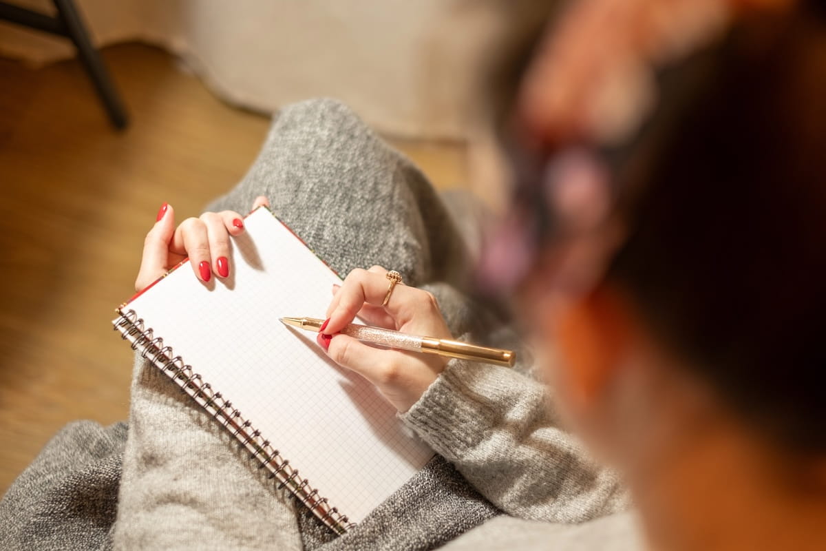 A high-angle, over-the-shoulder shot of a woman with her hair in a bun, wearing a grey knit sweater and red nail polish. She is sitting and writing in a spiral-bound notebook with grid paper using a gold-and-silver pen. The focus is on her hands and the notebook, while the back of her head is softly blurred in the foreground, creating an intimate scene of focused study or journaling.