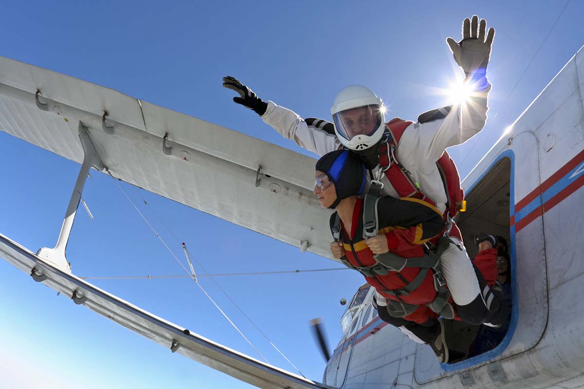 Tandem skydivers exit a small airplane under a clear blue sky. An instructor in a white helmet and jumpsuit is strapped to a participant wearing a red jumpsuit and goggles, with one hand raised as they begin their freefall. Sunlight shines behind them near the aircraft door.