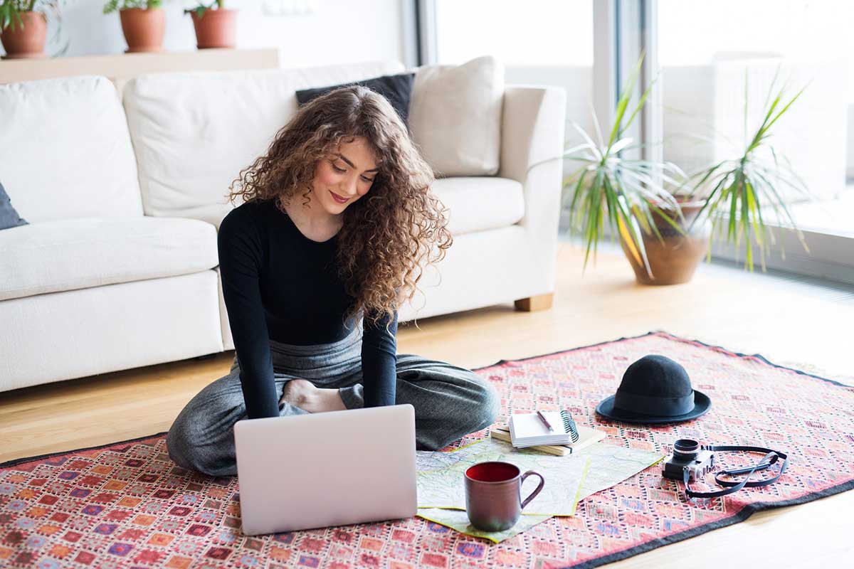 Woman sat on the carpet planning a summer vacation on her laptop.