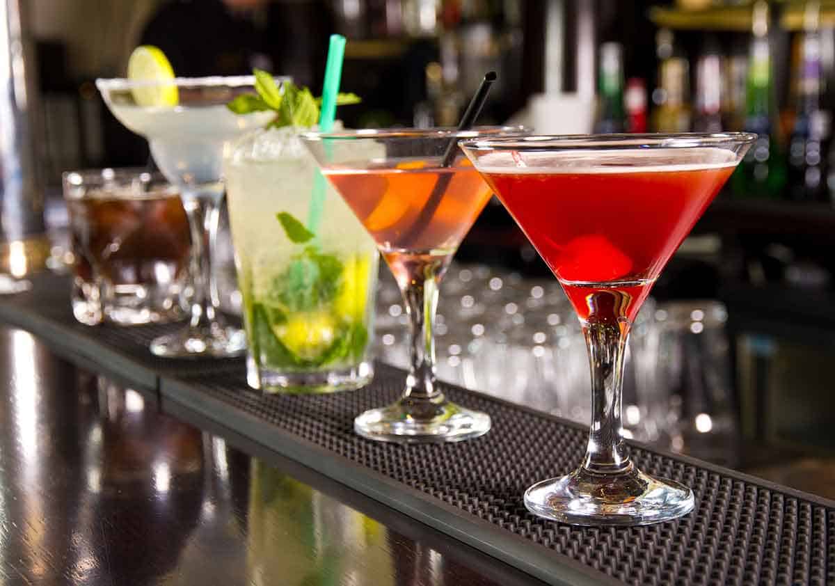Assorted colorful cocktails arranged on a bar counter, including red, pink, green, and clear drinks in martini and highball glasses. The drinks are garnished with items like lime, mint, and cherries, with a blurred background of bottles and glassware.