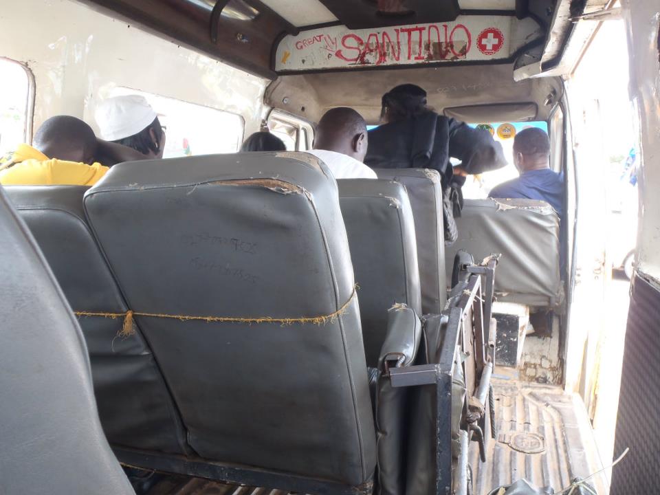 An interior, eye-level view from the back of a trotro (minibus) in Ghana. The cabin features several rows of worn, grey vinyl seats with visible tears and some secured with yellow twine. Five passengers are visible from the back, including a man in a yellow shirt and a white kufi cap on the left, and a conductor standing near the open sliding door on the right. Above the windshield, hand-painted red and black text reads "GREAT SANTINO" next to a red medical cross symbol. The van has a weathered white interior and a ribbed metal floor.