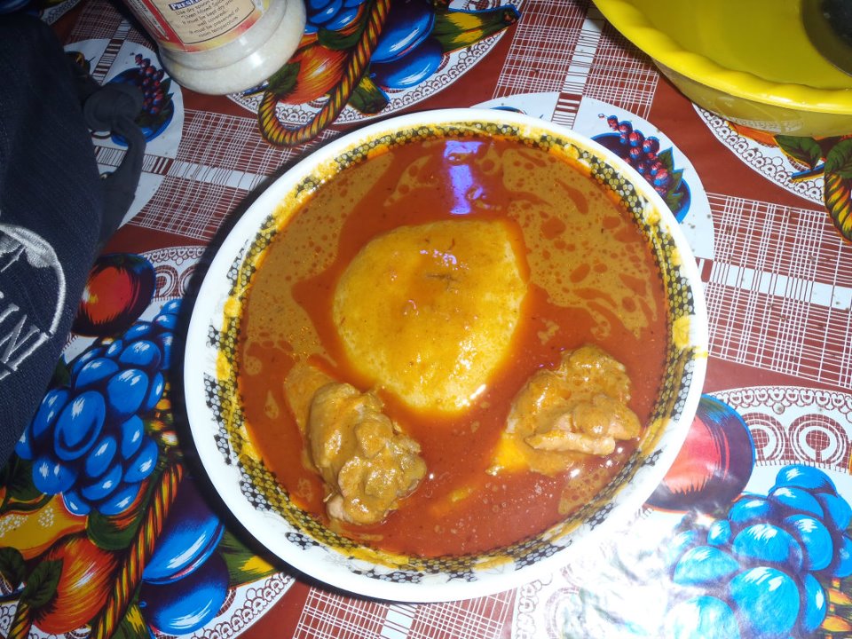 A top-down shot of a traditional Ghanaian meal featuring a large, smooth ball of pale yellow fufu centered in a bowl of vibrant, orange-red groundnut (peanut) soup. Two pieces of bone-in chicken are visible in the rich broth. The bowl is set on a colorful tablecloth with a bright floral pattern of blue grapes and red fruit. A small portion of a yellow bowl is visible in the upper right corner.