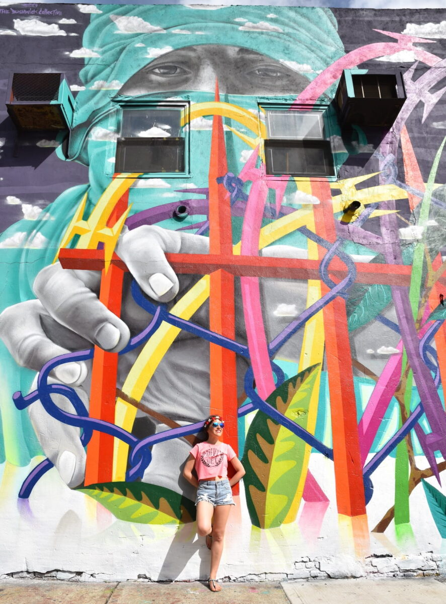 A traveler posing in front of a collaborative street art mural by Chilean artists Dasic Fernรกndez and Cekis at the Bushwick Collective on Troutman Street in Brooklyn, NYC.