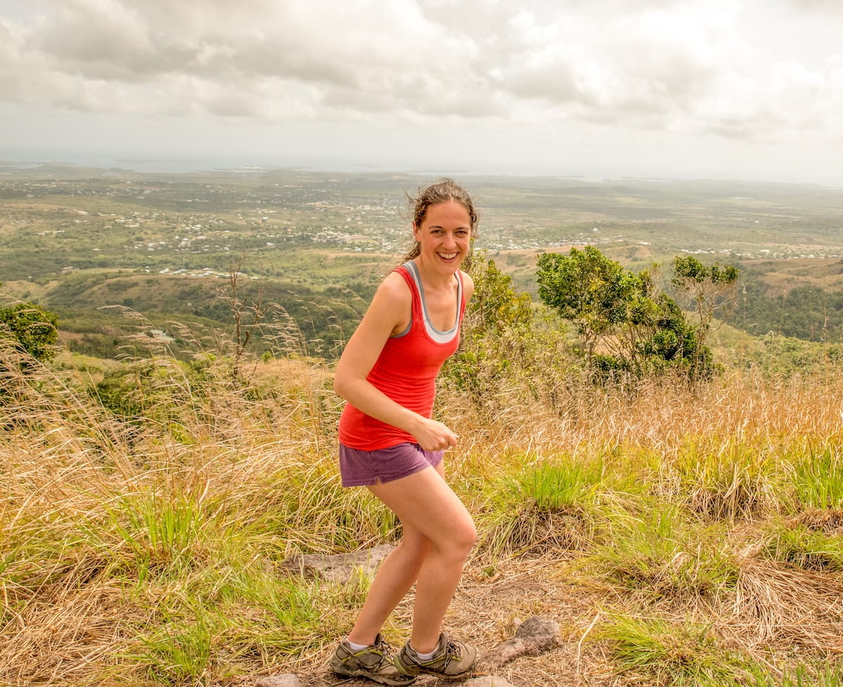 female hiker in a red tank top smiling after reaching the top of Signal Hill with an expansive view over Antigua
