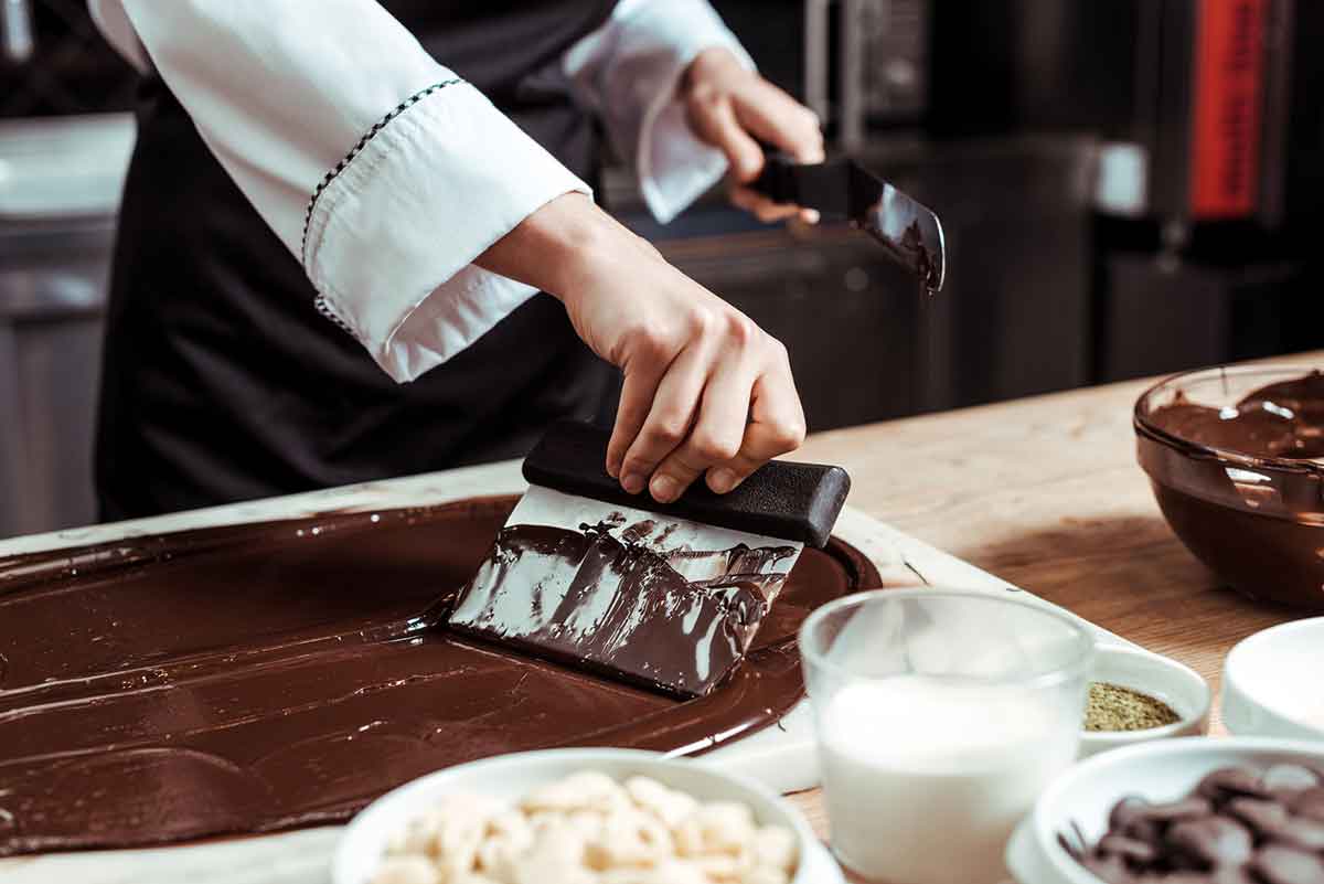 Cropped view of chocolatier holding cake scraper near melted dark chocolate and milk.
