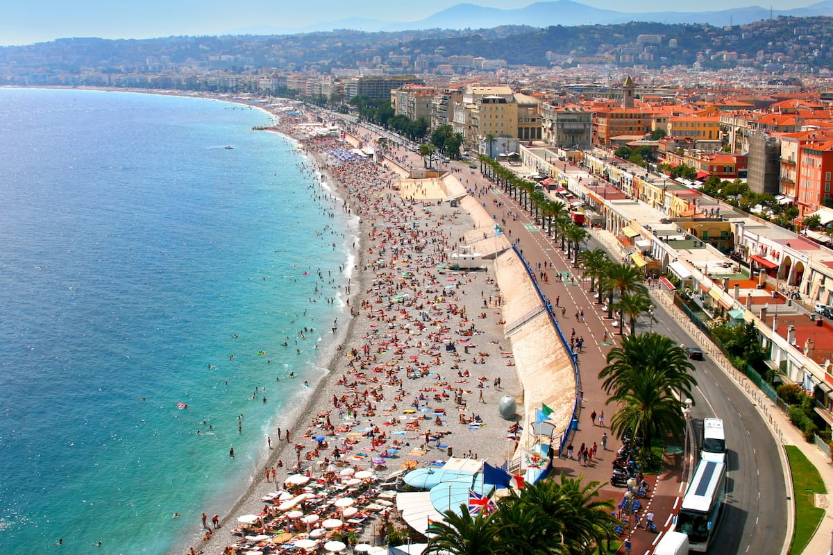 An elevated, wide-angle view of the crowded pebble beach in Nice, France, featuring turquoise Mediterranean waters on the left and the Promenade des Anglais lined with palm trees and historic coastal buildings on the right under a clear blue sky.