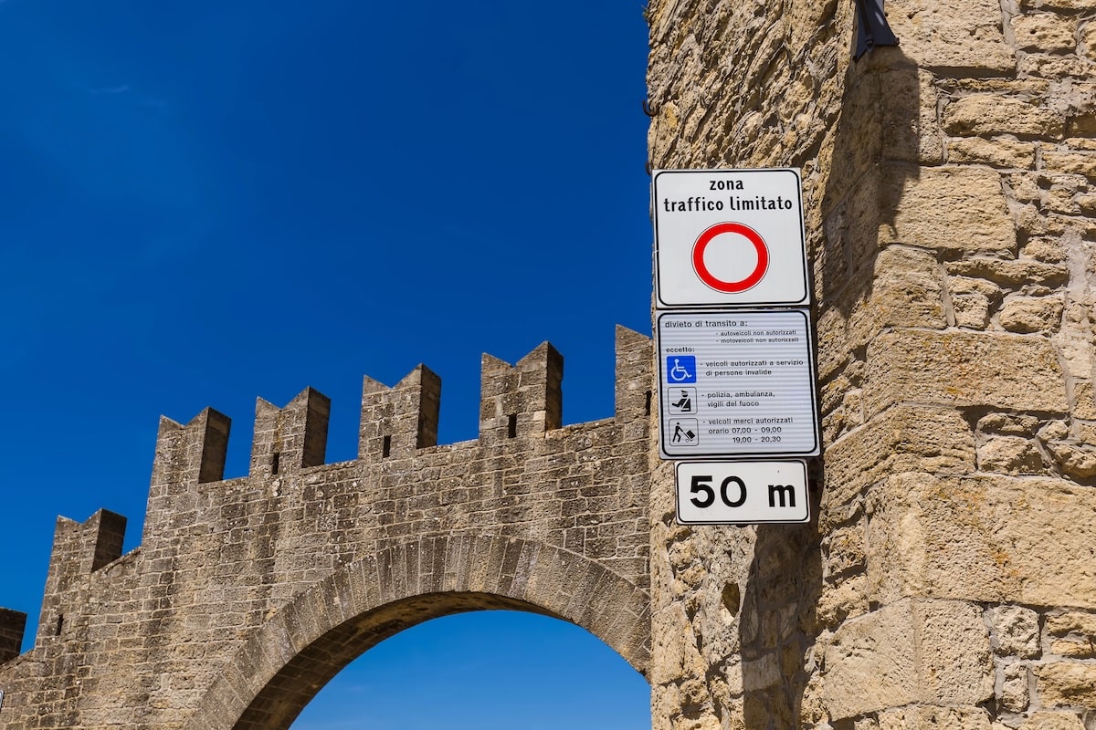 A vertical shot of a set of Italian traffic signs mounted on an ancient, light-brown stone wall with a crenelated top and an archway. The top sign reads "zona traffico limitato" (ZTL) above a red circle, indicating a restricted driving zone. A smaller sign below lists exceptions for disabled parking, emergency vehicles, and delivery times, followed by a bottom sign that reads "50 m." The scene is set against a deep, cloudless blue sky.