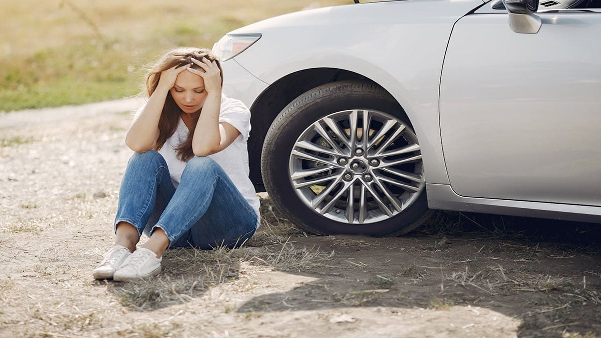 Young woman sitting on the ground next to a car with a flat tire, holding her head in frustration. She appears distressed, highlighting a common travel horror story scenario. The car is parked on a dirt road, emphasizing the challenging and unexpected situations that can occur while traveling.