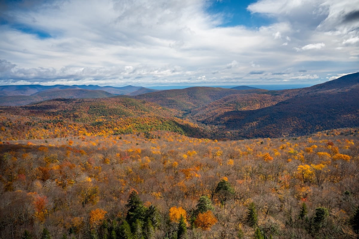 A wide panoramic landscape view looking out from Giant Ledge in the Catskills during peak October autumn foliage. Rolling mountain ridges and valleys stretch across the background under a dynamic sky filled with wispy clouds and patches of blue. Below, a vast forest canopy displays a rich blend of rust, orange, golden yellow, and deep evergreen foliage, with dappled sunlight creating soft shadows across the valley floor.