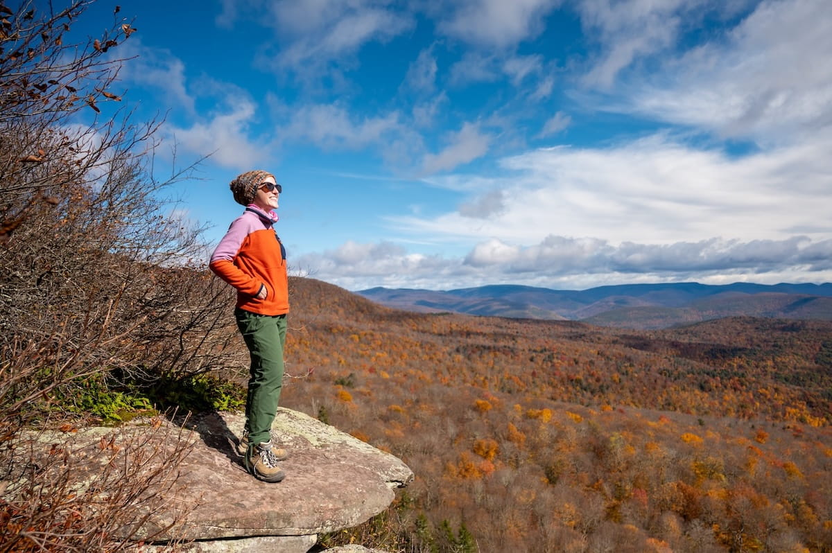 A side-profile full-body shot of a female hiker standing on a flat stone ledge at a viewpoint along the Giant Ledge Trail in the Catskills. She wears sunglasses, a knit beanie, an orange and pink long-sleeve top, green hiking pants, and brown hiking boots, smiling as she looks out over the landscape with her hands in her pockets. Below the ledge, a vast forest stretches across rolling mountains blanketed in rich orange, yellow, and red autumn colors beneath a blue sky filled with wispy white clouds.