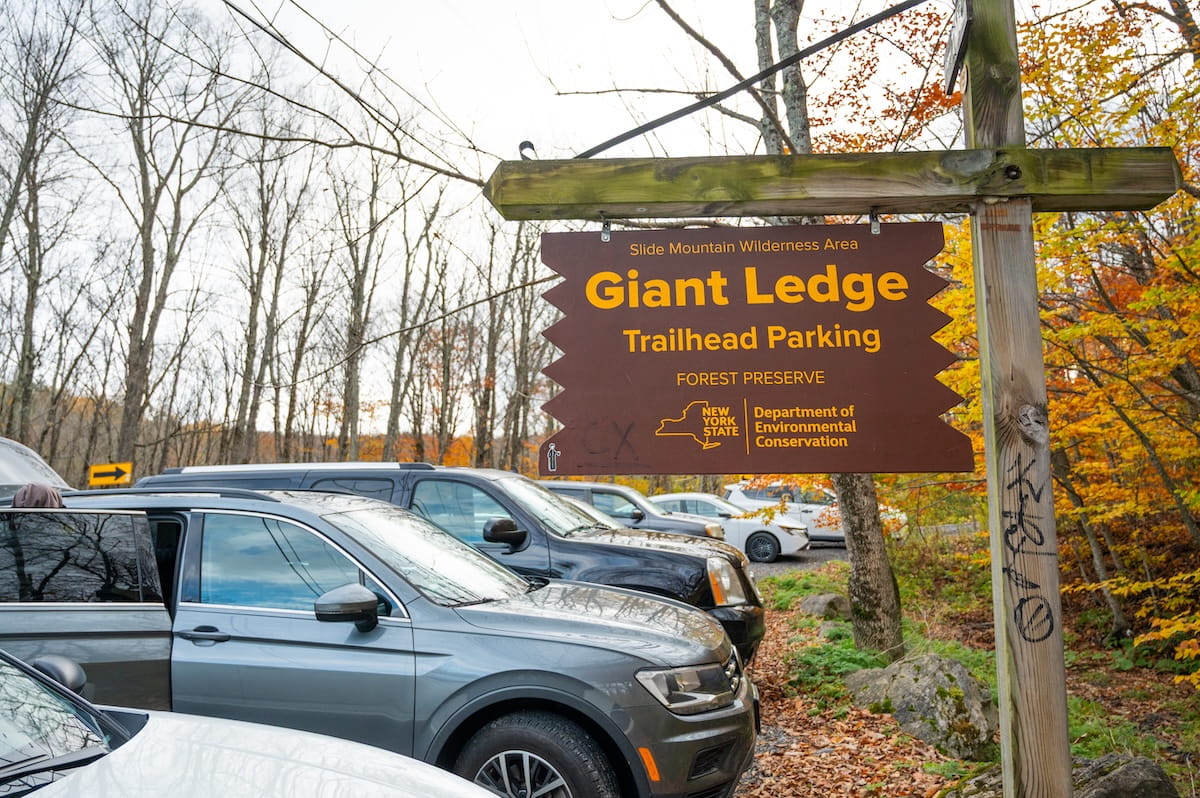 A close-up shot of a dark brown wooden state sign with yellow text reading "Slide Mountain Wilderness Area - Giant Ledge Trailhead Parking - Forest Preserve" and the New York State Department of Environmental Conservation logo. The sign is mounted on a thick wooden post in front of a parking lot filled with parked cars, surrounded by autumn trees showing bare branches and colorful fall foliage.