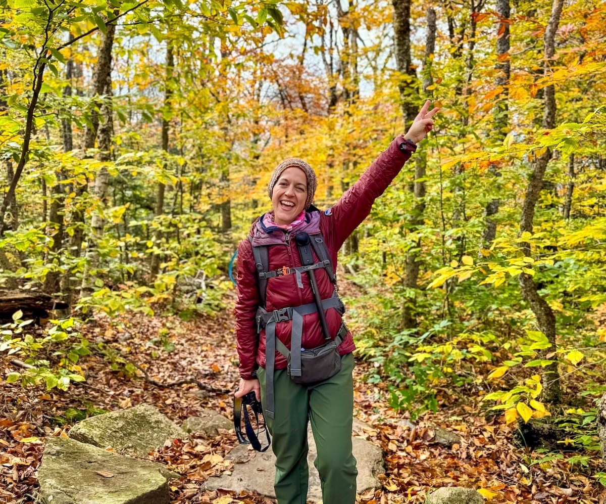 A medium shot of a smiling female hiker flashing a peace sign on the Giant Ledge Trail in the Catskills. She is wearing a brown knit beanie, a maroon puffer jacket, a grey hiking backpack with chest straps, and green pants, holding a camera strap in her other hand. She stands on a leaf-strewn, rocky forest path surrounded by vibrant yellow and green autumn foliage.