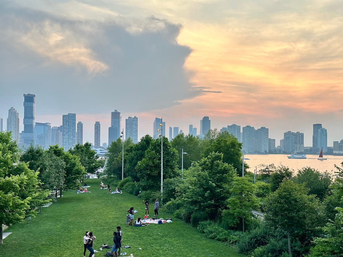 A high-angle view of a lush green lawn at City Vineyard in NYC during sunset. Groups of people are scattered across the grass, lounging and enjoying picnics. A thick line of vibrant green trees separates the park from the Hudson River, where a few boats are visible. In the background, the Jersey City skyline stands beneath a dramatic sky filled with large, dark grey clouds contrasting against a soft orange and yellow glow from the setting sun.