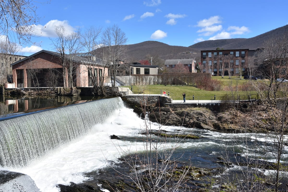 A wide landscape shot of Fishkill Overlook Falls in Beacon, New York. Water cascades over a wide, low dam spillway into a rocky stream with white rushing foam in the foreground. On the opposite bank sits an open brick building, manicured green lawns, and historic brick mill buildings, with rolled mountains rising in the background under a sunny blue sky dotted with light clouds.