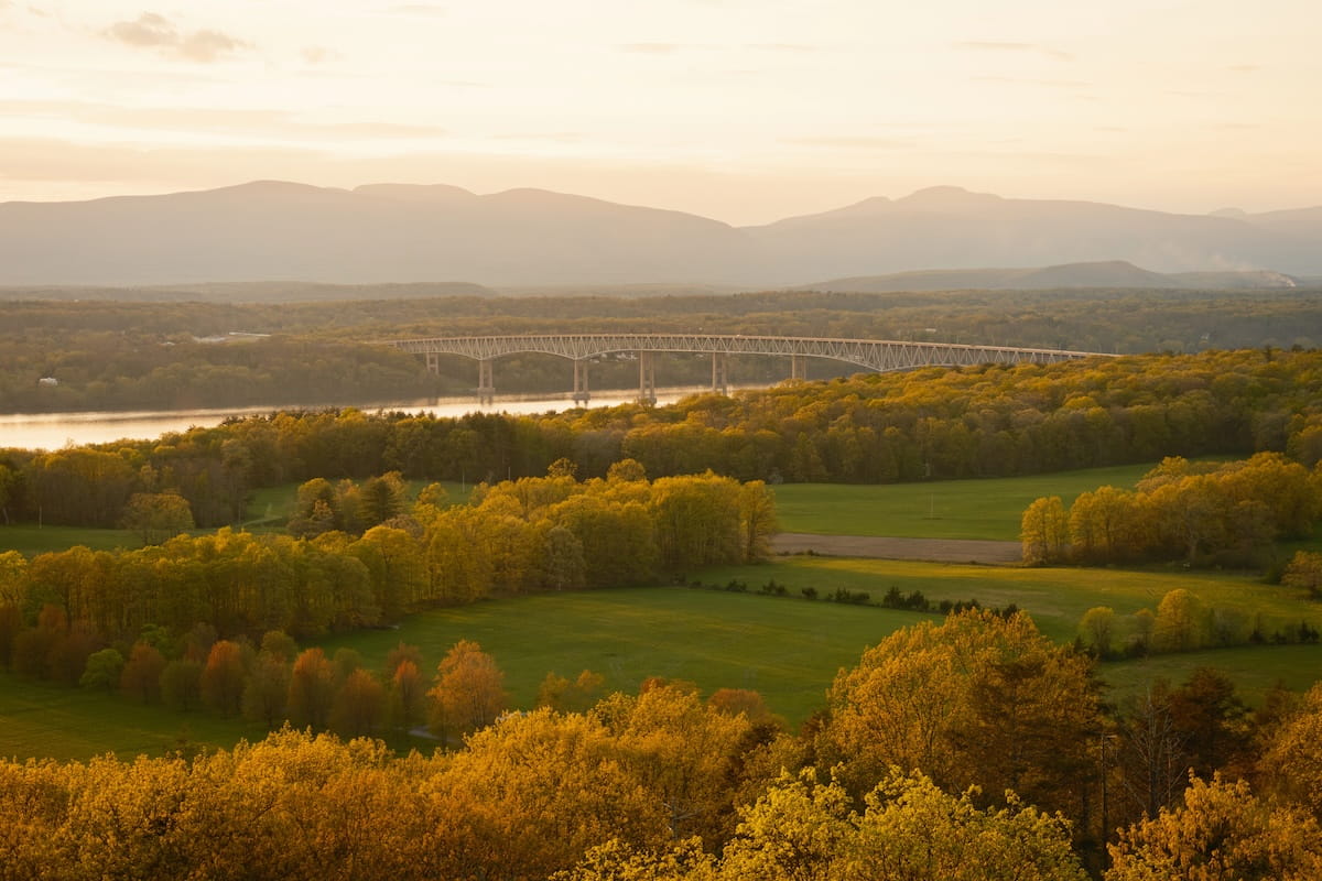 A high-angle landscape shot capturing a golden sunset view near Rhinebeck, New York. Warm, golden sunlight illuminates lush green fields and dense, yellow-tinged treetops in the foreground and midground. In the background, the Hudson River flows past, spanned by a long continuous truss bridge, with rolling mountain ridges stretching under a soft, hazy sky.
