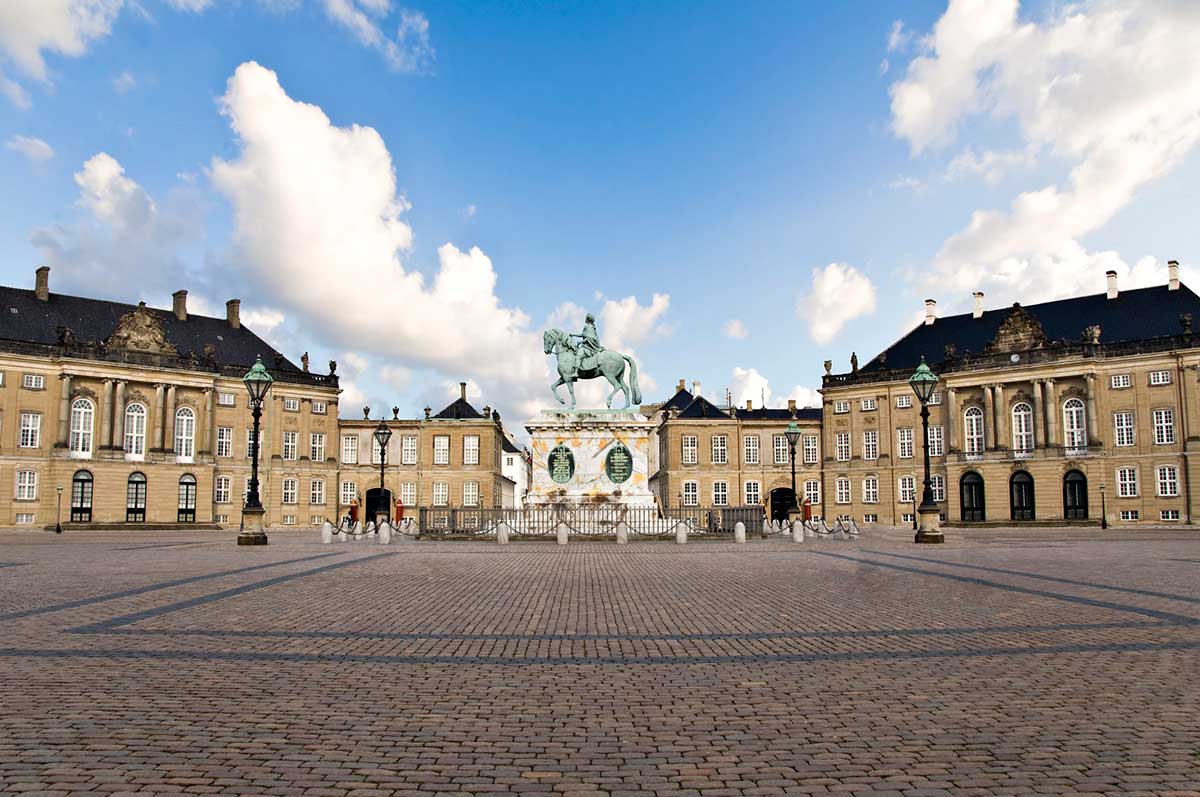 A wide view of Amalienborg Palace in Copenhagen showing the symmetrical palace buildings surrounding a cobblestone square, with an equestrian statue at the center under a partly cloudy sky.