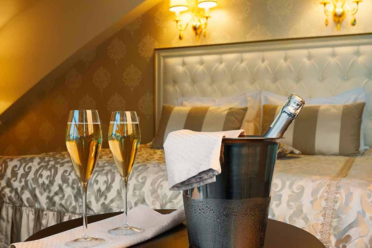 A luxurious hotel room scene featuring a bottle of champagne chilled in an ice bucket with a cloth draped over it, two champagne glasses on a table, and a neatly made bed with a tufted headboard and warm ambient lighting in the background.