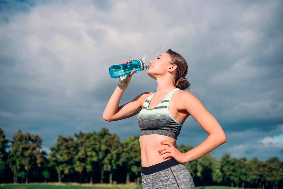Woman in athletic clothing standing outdoors, drinking from a water bottle with one hand on their hip. Trees and a cloudy sky are visible in the background.