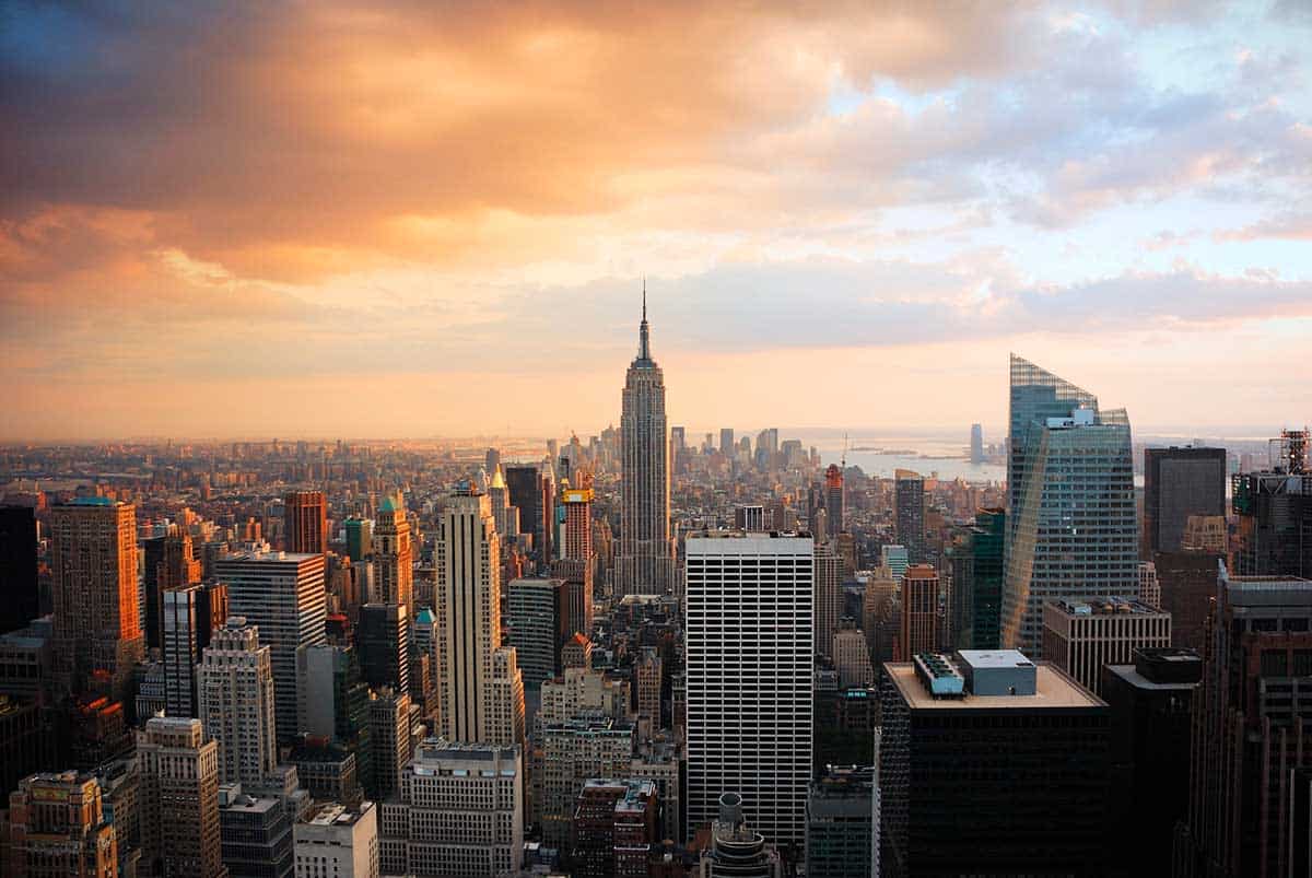A panoramic view of the Manhattan skyline during sunset, featuring the Empire State Building at the center surrounded by tall skyscrapers, with warm golden light reflecting off the buildings and the harbor visible in the distance.