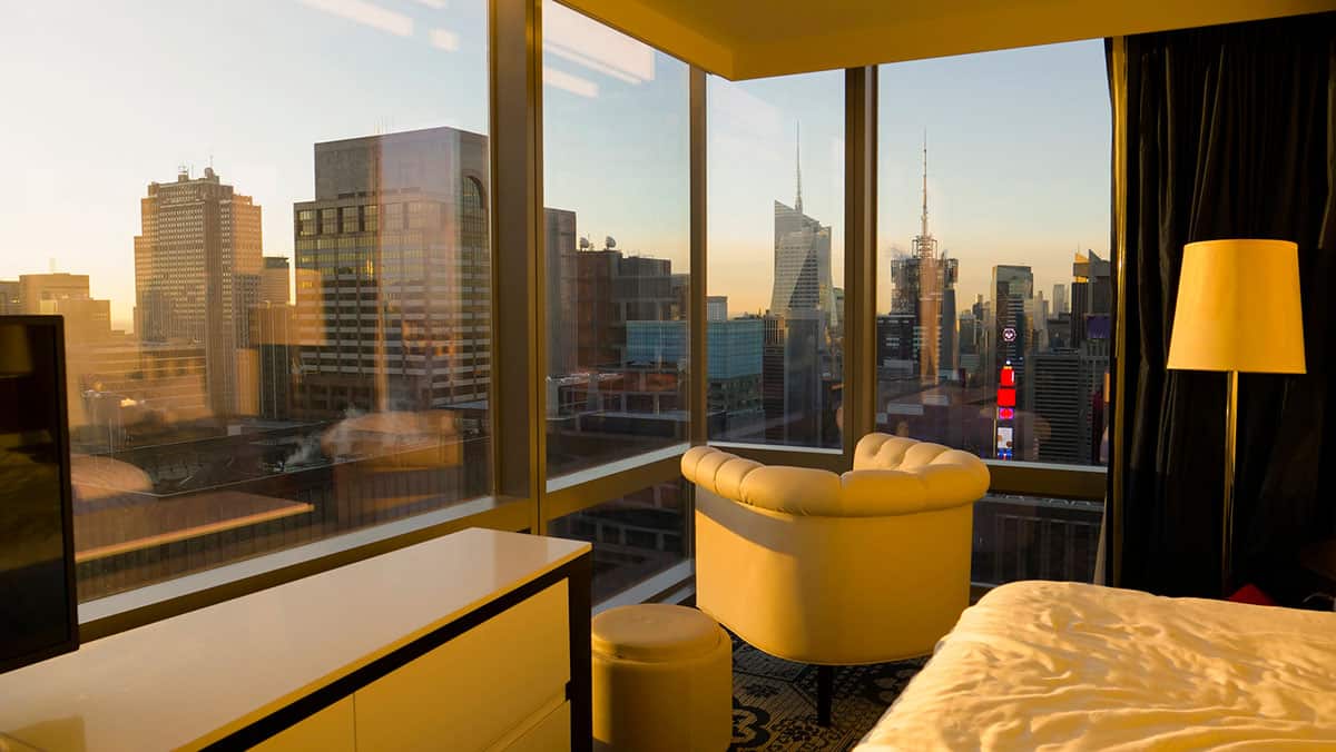 A stylish NYC hotel room featuring floor-to-ceiling windows, a bed and armchair in the foreground, and a sweeping view of the city skyline illuminated by warm sunset light.