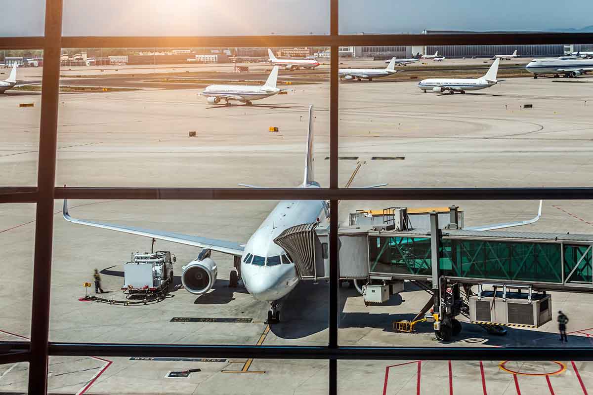 Airplane parked at an airport gate with a jet bridge, viewed through terminal windows, with other planes on the runway in the background.