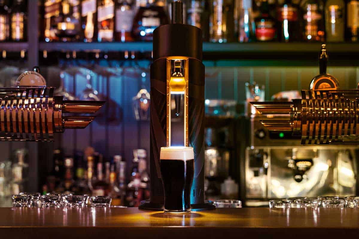 A pint of dark stout with a creamy head sits on a bar counter beneath a beer tap, with bottles and bar equipment in the background.