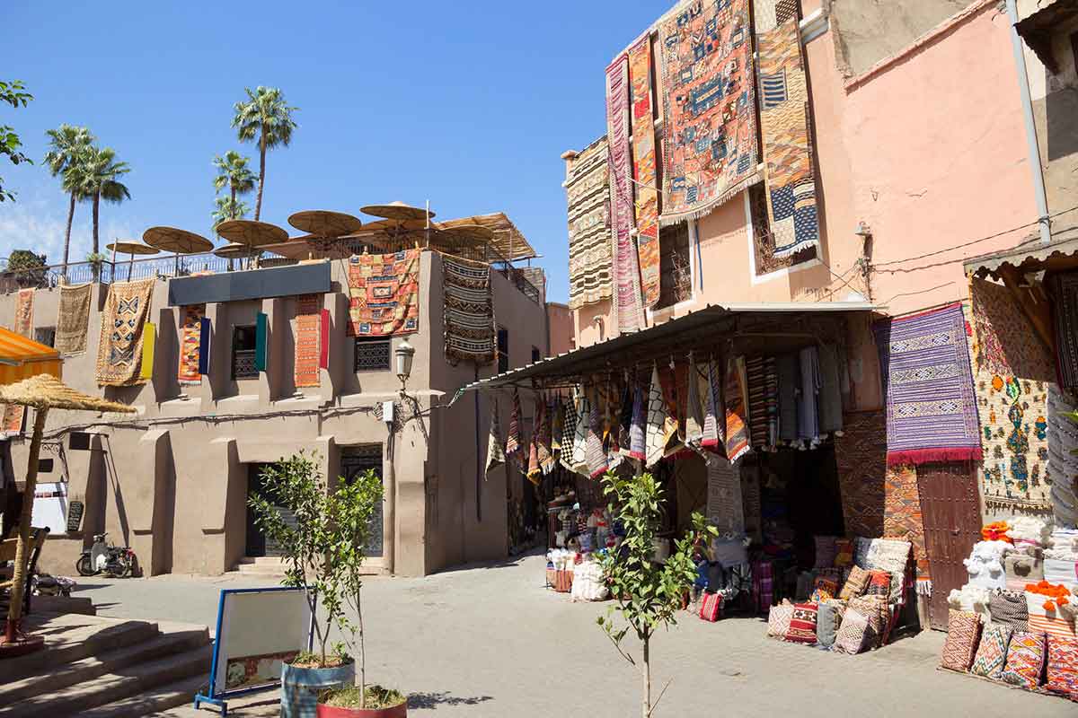 Traditional souk in Marrakech with colorful woven rugs and textiles hanging from market stalls and buildings under a clear blue sky.