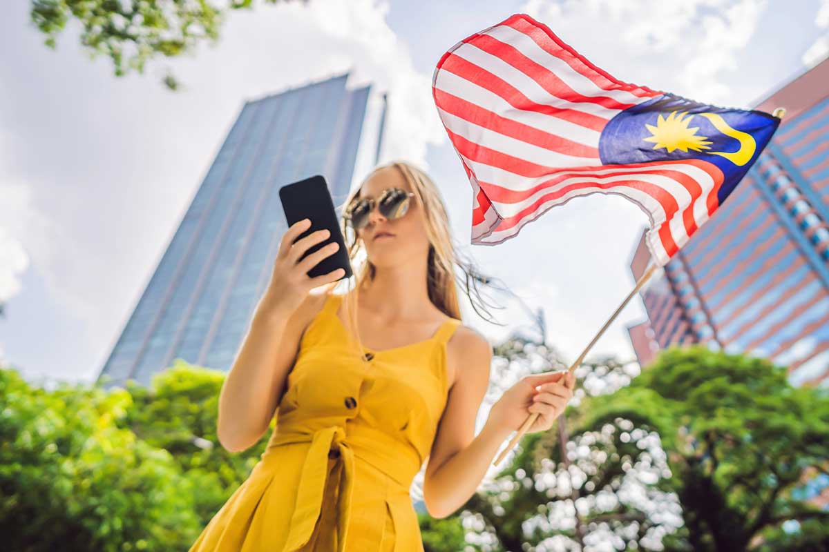 A woman in a yellow dress stands outdoors in a city, holding a smartphone and a Malaysian flag, with tall buildings and trees in the background.