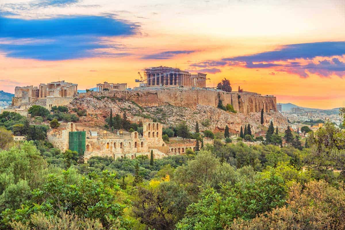 A panoramic scene of the Acropolis in Athens at sunset, featuring the Parthenon and surrounding ancient structures illuminated by warm golden light, with lush green trees in the foreground and a dramatic sky of orange, pink, and blue hues overhead.