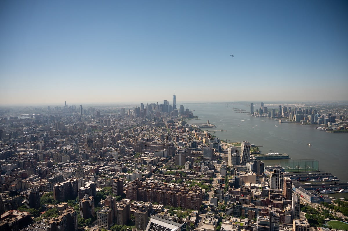 A high-angle, wide-reaching panoramic view of the Manhattan skyline from the Edge observation deck, looking south toward One World Trade Center and the Hudson River under a clear blue sky.