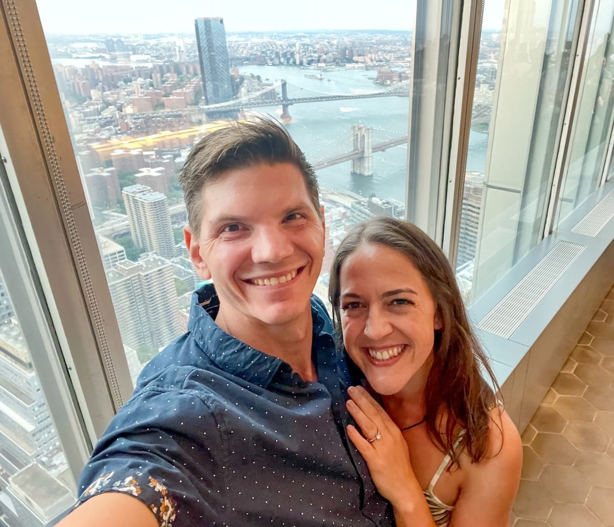A smiling couple takes a selfie inside the Manhatta rooftop restaurant, with floor-to-ceiling windows behind them showcasing a high-altitude, panoramic view of the East River, the Brooklyn Bridge, and the Manhattan Bridge stretching into Brooklyn.