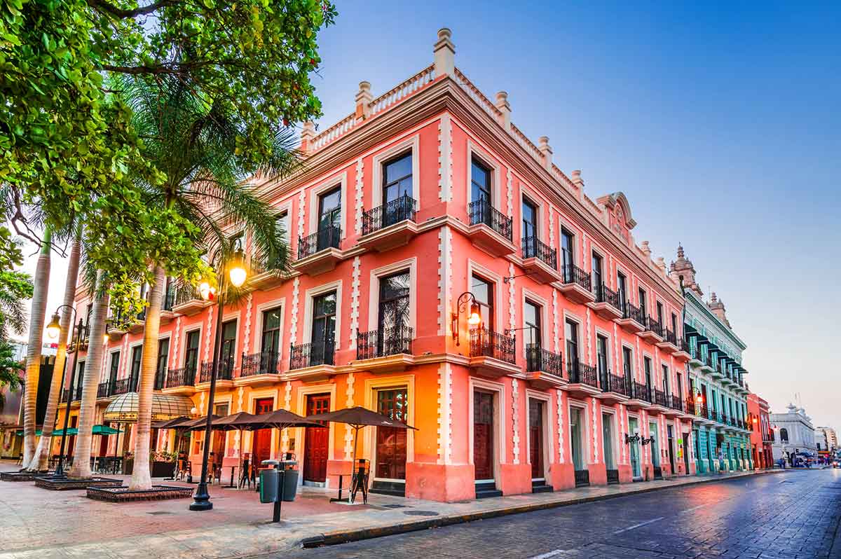 Colorful colonial building on a street corner in Mรฉrida, Mexico, with a pink faรงade, white trim, wrought-iron balconies, palm trees, and warm streetlights under a clear blue sky.