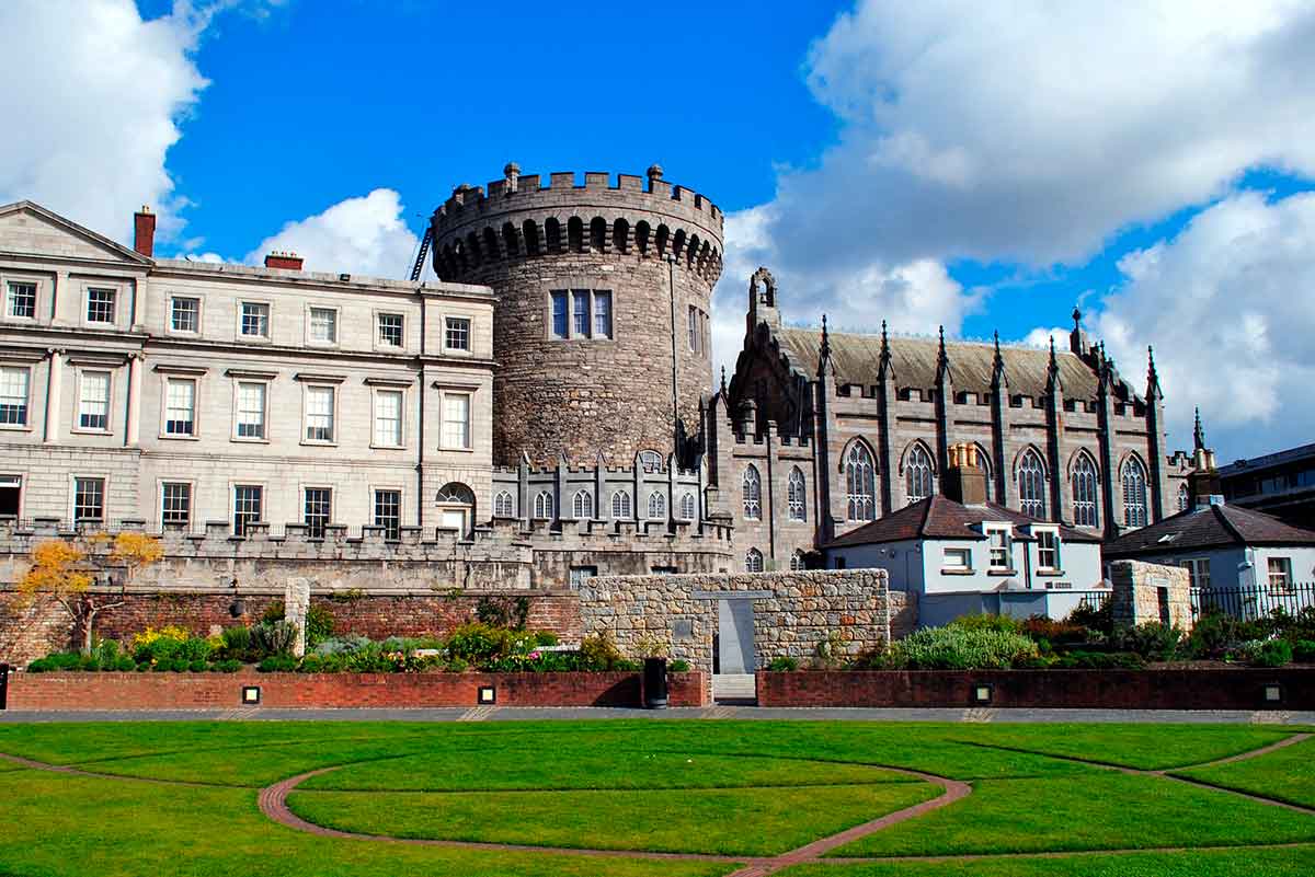 Dublin Castle with its round stone tower and historic buildings, set against a blue sky with clouds and a green lawn in the foreground.