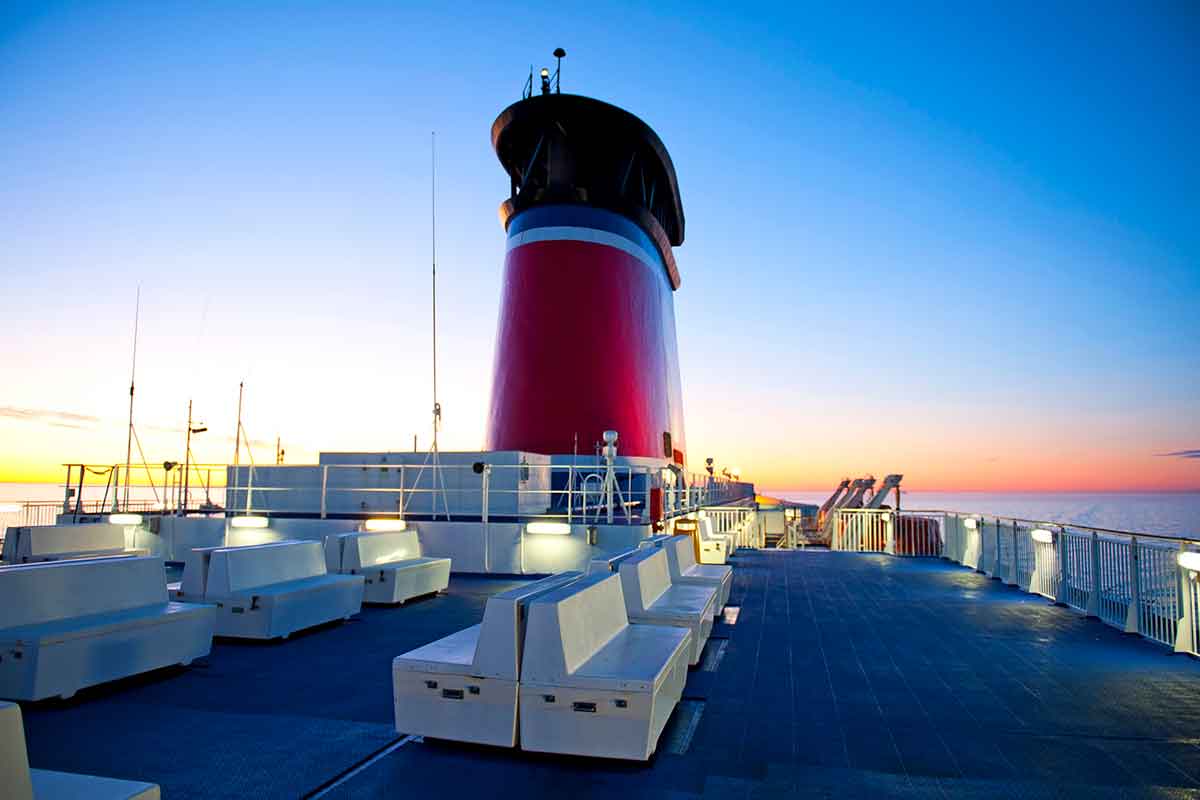 An empty ferry deck at sunset with white benches and railings, featuring a large red smokestack and the ocean visible in the background.