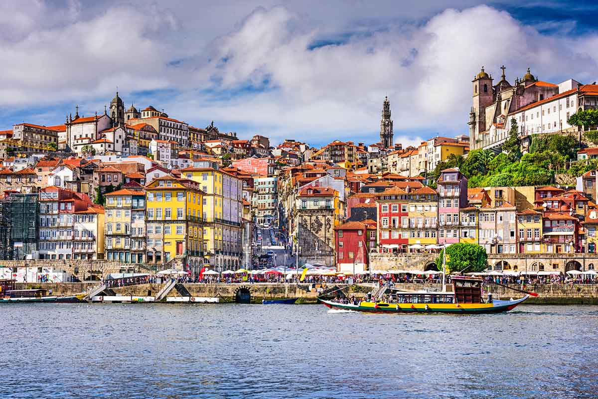 The colorful skyline of Porto, Portugal, with historic buildings stacked along the hillside, a riverside promenade, and a traditional boat sailing on the Douro River.