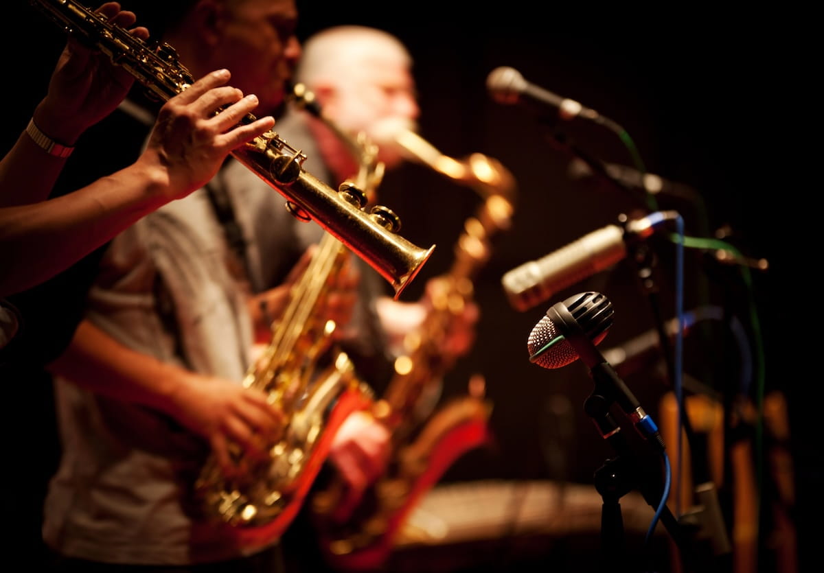 A side-profile, close-up shot of three musicians playing saxophones during a live performance in a dimly lit jazz club. The musician in the immediate foreground is in sharp focus, showing only their hands and the golden, reflective brass of the saxophone. The other two musicians and their instruments are blurred in the background. Several vintage-style microphones on stands are positioned in front of the players, all illuminated by warm, dramatic stage lighting against a dark backdrop.