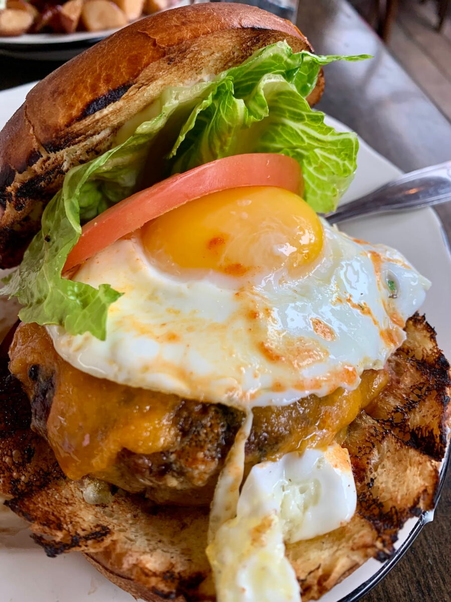 A mouth-watering close-up of a brunch burger on a toasted brioche bun. The burger features a thick, juicy beef patty melted with cheddar cheese and topped with a perfectly cooked sunny-side-up egg with a bright yellow yolk. A crisp green lettuce leaf and a fresh red tomato slice are tucked under the top bun. The burger is served on a white plate, with a side of golden-brown roasted potatoes partially visible in the blurred background.