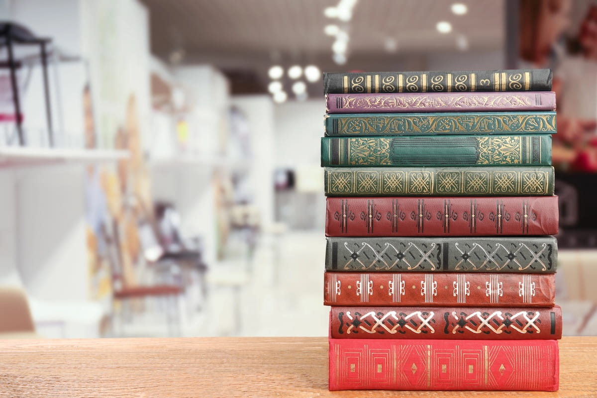A close-up, eye-level shot of a tall stack of ten vintage-style hardcover books resting on a light-colored wooden table. The book spines feature a variety of colorsโincluding deep red, forest green, navy blue, and charcoal greyโdecorated with intricate gold-leaf patterns and geometric designs. The background is a soft-focus, brightly lit interior of a bookstore or library, with blurred shelves and warm overhead lighting creating a clean and inviting atmosphere.
