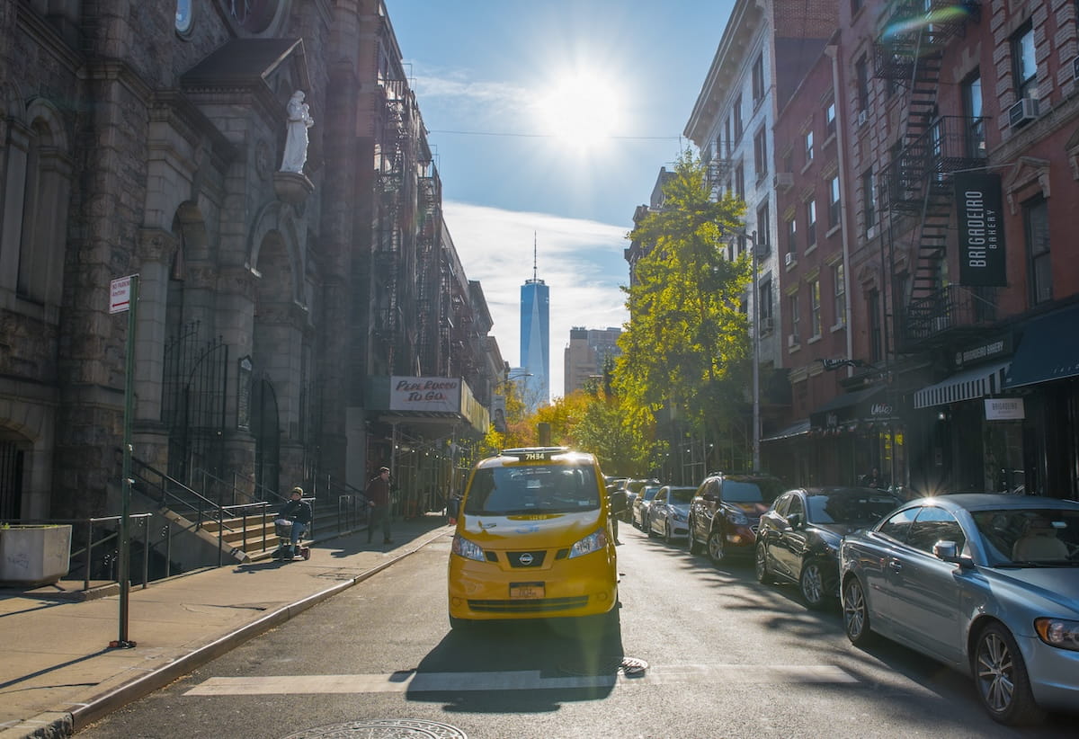A ground-level street view looking south down Sullivan Street in the West Village, NYC, on a bright, sunny day. A yellow taxi dominates the foreground, driving toward the viewer. In the distance, the One World Trade Center tower is framed perfectly by the canyon of buildings and autumn trees. To the left is the stone facade of a church featuring a white statue of a saint, and to the right is the red-brick exterior of Brigadeiro Bakery. The sun glows brightly at the top of the frame, casting long shadows across the asphalt.