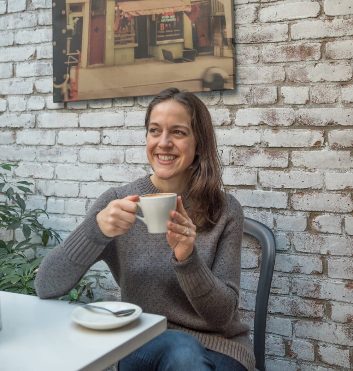 A medium shot of a smiling woman with long brown hair sitting at a white cafe table inside Pasticceria Rocco in the West Village, NYC. She is wearing a grey mock-neck sweater and holding a white ceramic cup of coffee with both hands. She is looking off-camera to her right. Behind her is a rustic, white-painted brick wall adorned with a framed photograph of a vintage storefront. A green potted plant is partially visible to the left.