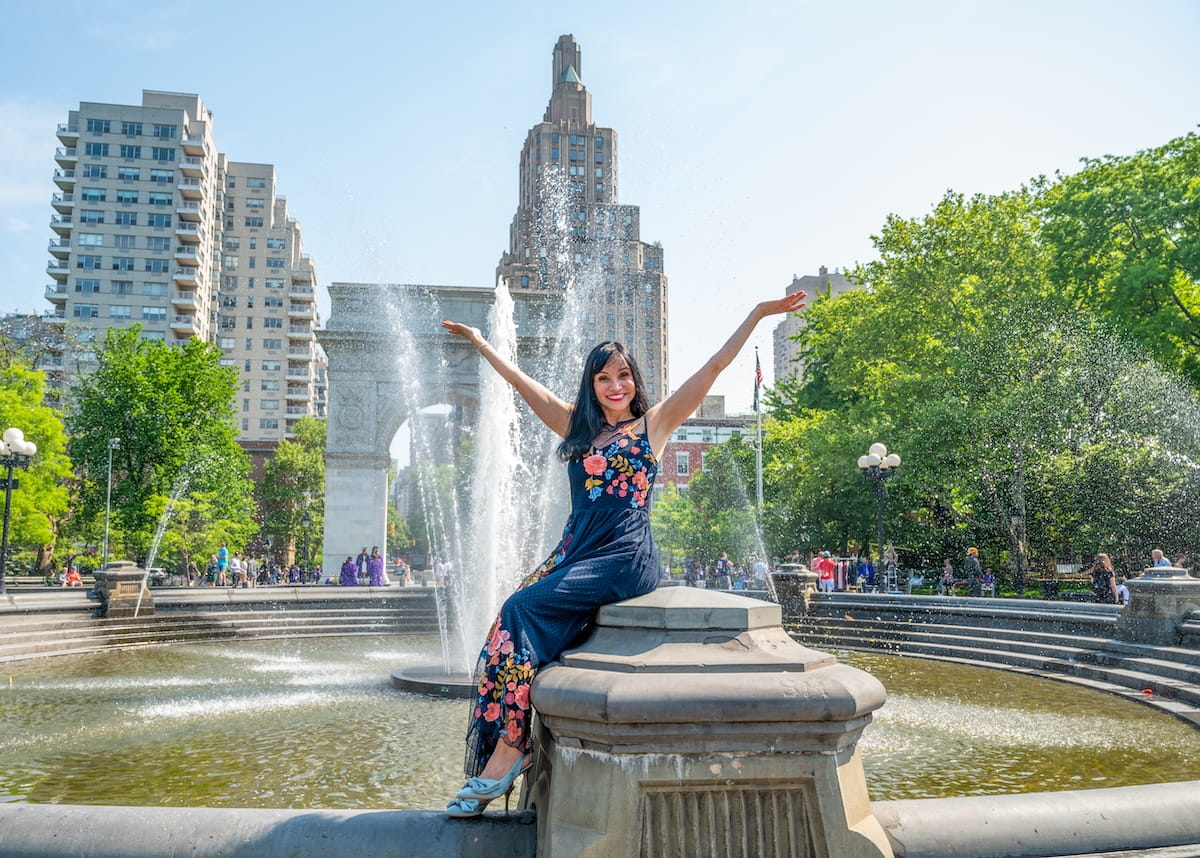 A medium-wide shot of a smiling woman with long dark hair sitting on the stone edge of the fountain in Washington Square Park, NYC. She is wearing a navy blue floral-embroidered maxi dress and has both arms raised joyfully toward the sky. In the background, water spray from the fountain frames the Washington Square Arch and the tall, historic residential buildings of Greenwich Village under a clear, bright sky.