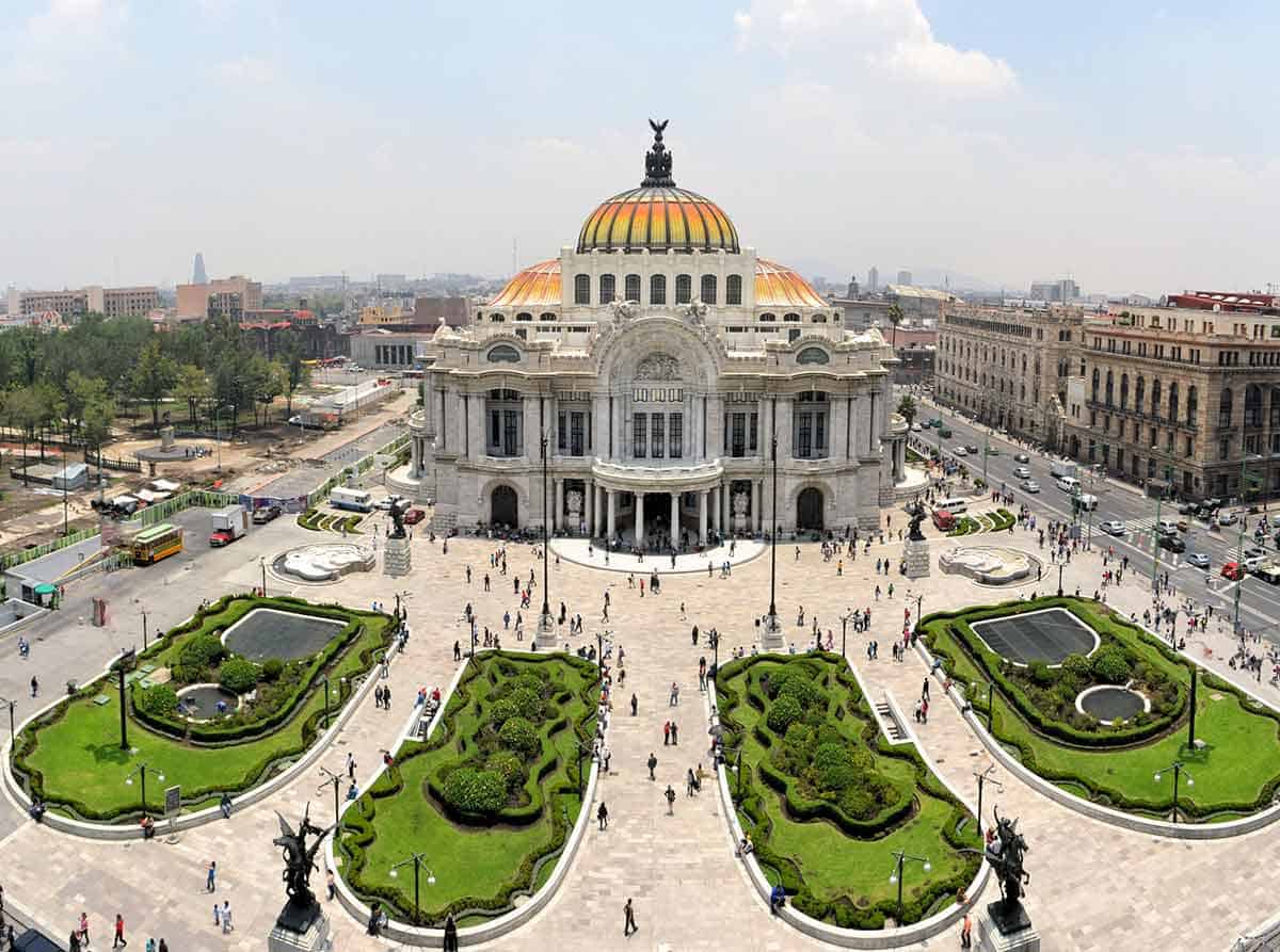 A wide aerial view of the Palacio de Bellas Artes in Mexico City, showing its white marble façade and colorful domed roof, framed by symmetrical gardens, statues, and a busy public square with pedestrians.