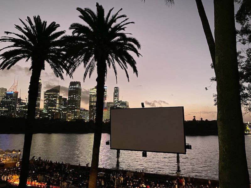 A big movie screen outside next to palm trees and the Sydney waterfront