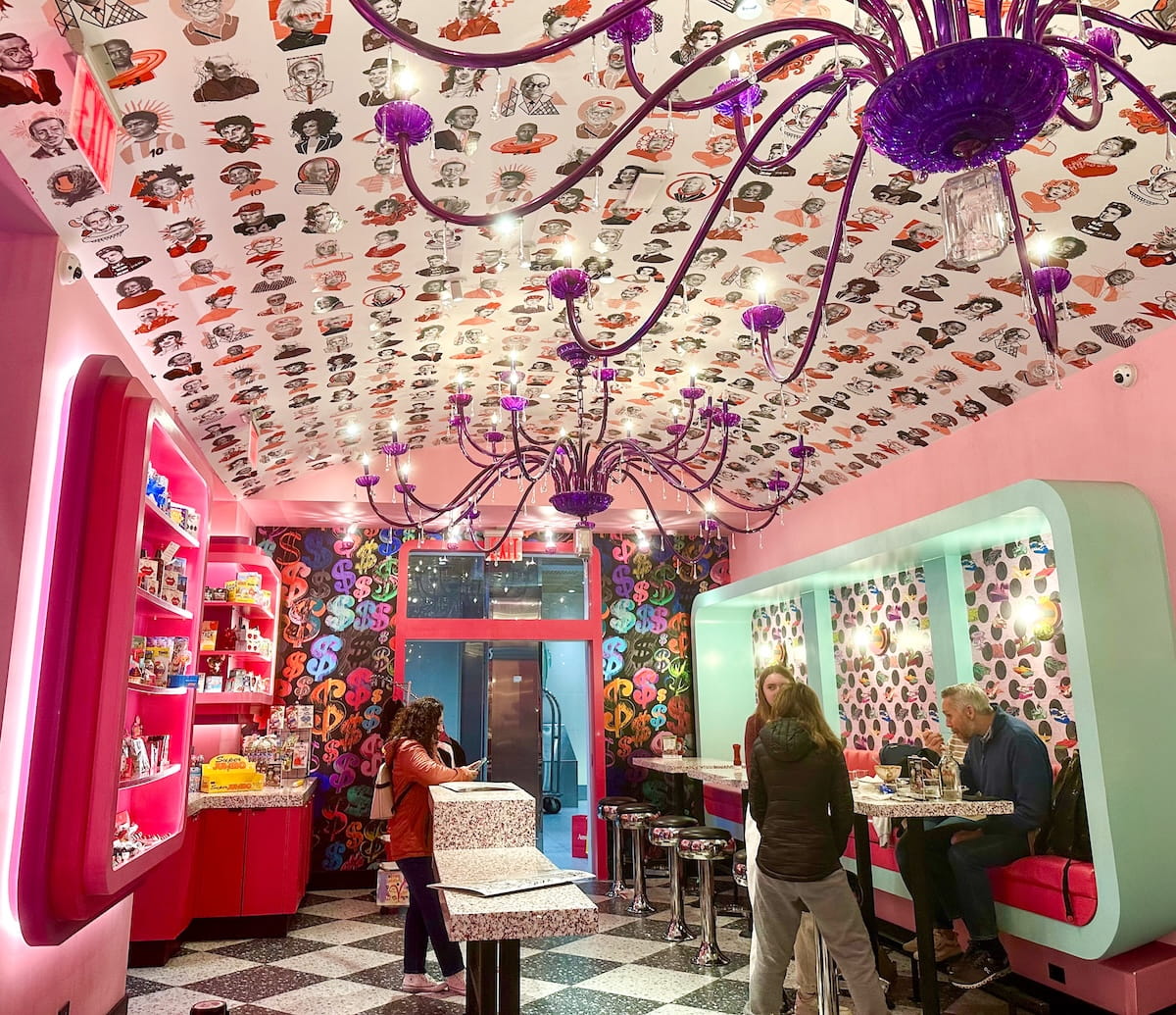 A wide-angle interior view of the whimsical Serendipity3 restaurant in NYC. The room features bubblegum-pink walls, a black-and-white checkered floor, and a ceiling covered in a pattern of colorful celebrity portraits. A large, ornate purple chandelier hangs from the center. On the right, diners sit in light-blue recessed booths, while pink shelving units filled with sweets line the left. In the background, a wall is covered in multicolored Warhol-style dollar sign wallpaper.