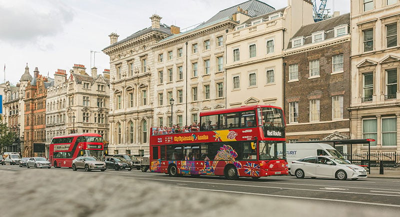 A red double decker bus driving down a street next to tall buildings.