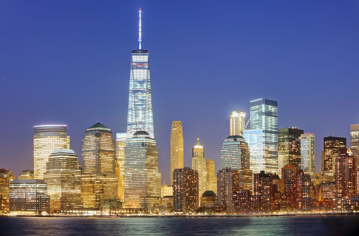An evening wide shot of the Lower Manhattan skyline across the Hudson River. One World Trade Center stands prominently in the center, its tall glass facade and white spire brightly illuminated against a deep blue twilight sky. Surrounding skyscrapers are densely packed and glowing with warm interior lights, casting soft reflections on the dark water in the foreground.