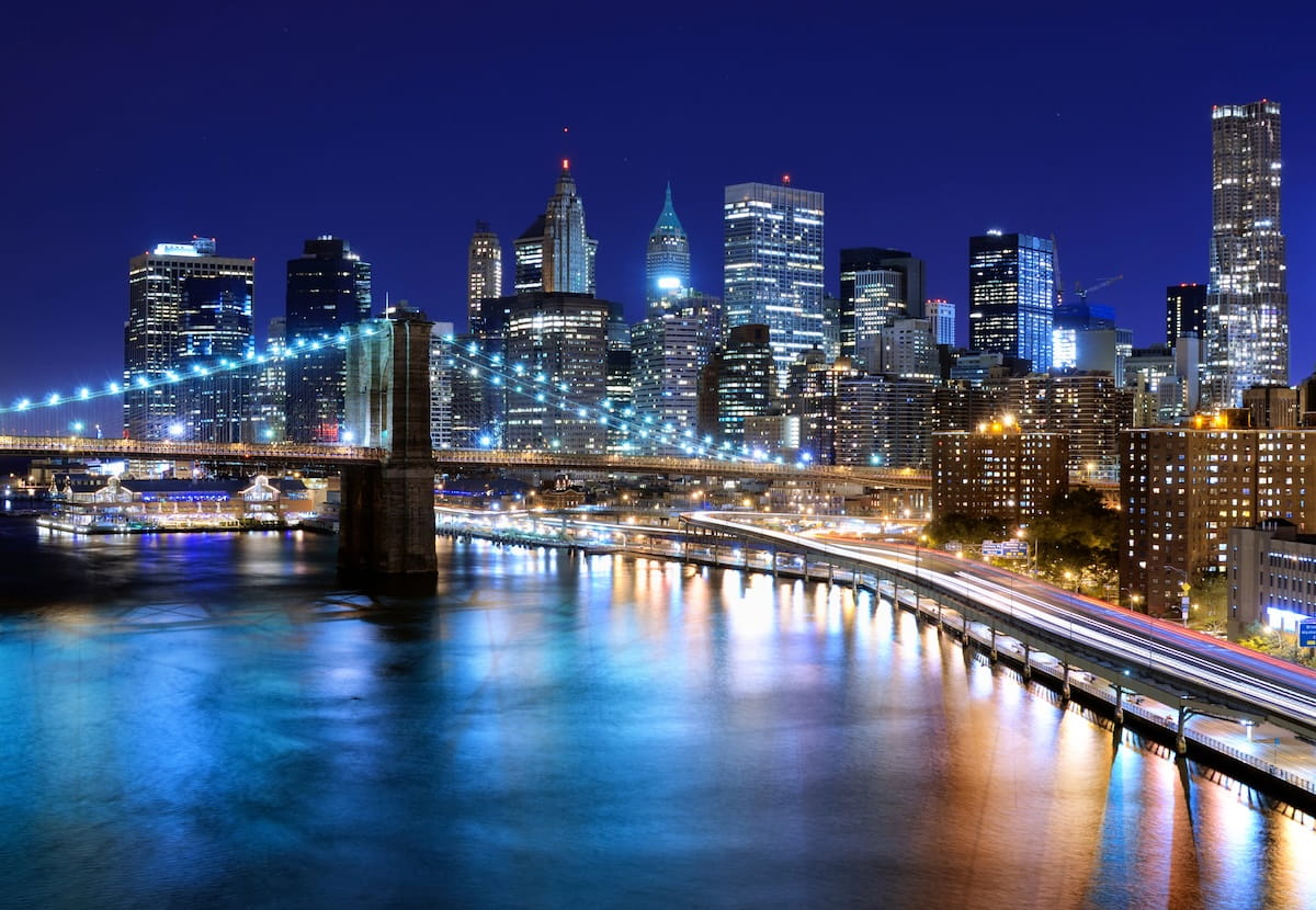 A high-angle, night-time view of the Lower Manhattan skyline and the Brooklyn Bridge stretching across the East River. The bridge crosses from the left, its stone tower and suspension cables illuminated by a string of bright white lights, while an elevated highway curves along the waterfront below, showing long-exposure light trails from traffic. The background features densely packed skyscrapers glowing with office lights against a dark blue night sky, casting vibrant blue, gold, and white reflections across the rippling river surface.