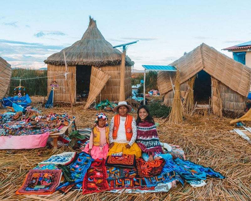 solo female traveler posing for a photo with the locals of the Uros Islands while sitting on totora reeds and handmade blankets 