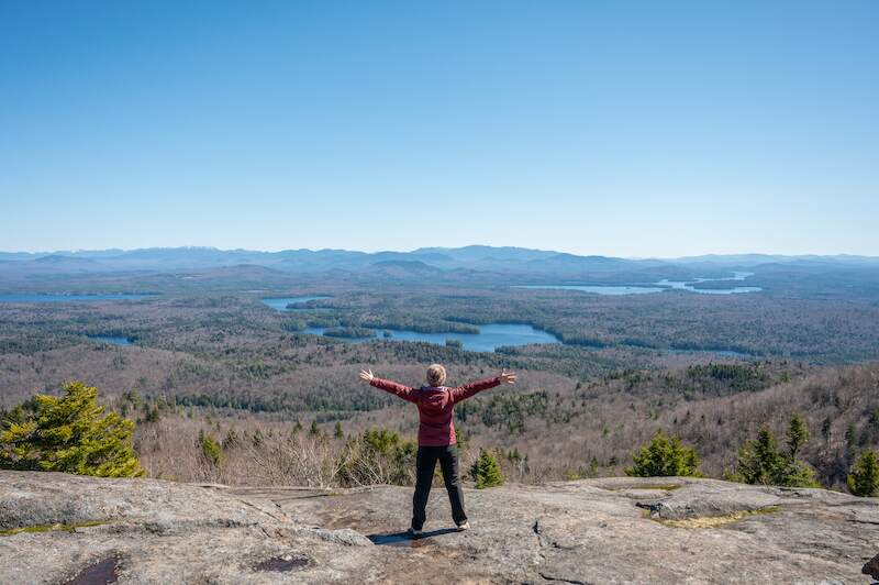 hiker posing at the top of Saint Regis Mountain Summit with views of the surrounding high peaks, lakes, and ponds in the Adirondacks Region of New York